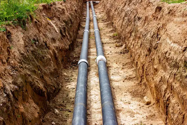 Two large black pipes, part of a sewer line installation, lay snugly in a narrow trench dug through the brown soil, surrounded by verdant grass.