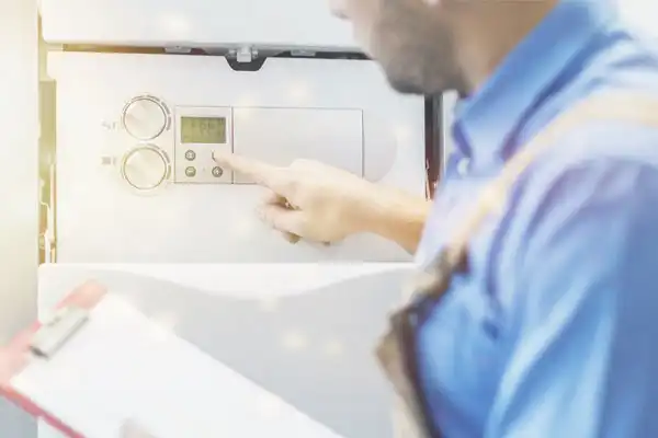 Person in blue shirt adjusts controls on a boiler, holding a clipboard, as part of a tankless water heater installation.