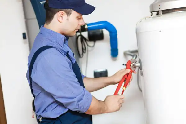 A person in a blue uniform and cap uses a wrench to perform water heater maintenance on a large white tank, with blue pipes visible in the background indoors.