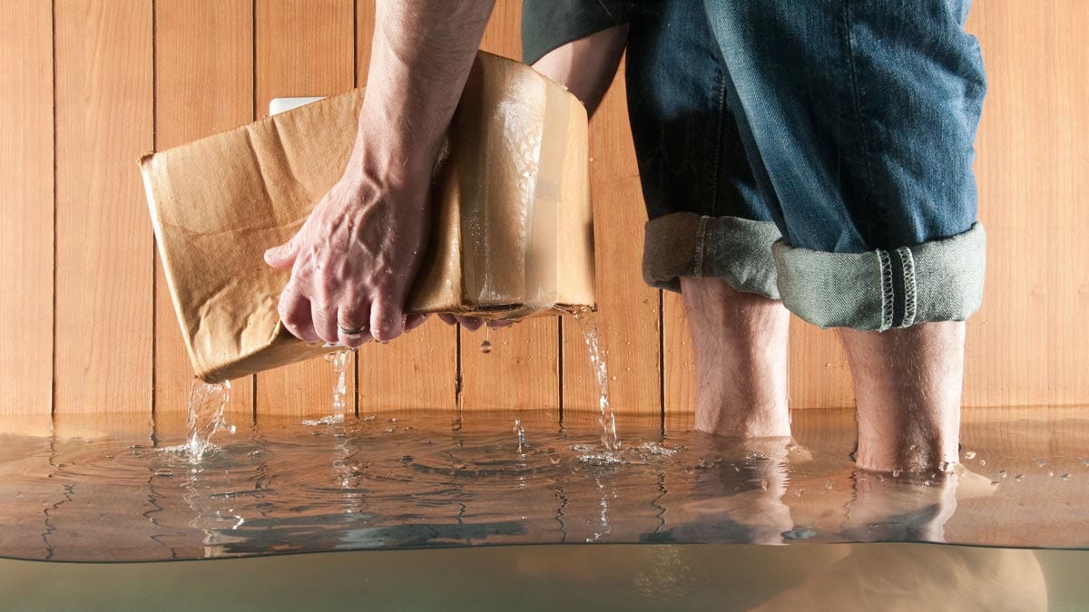 Person lifting a wet cardboard box from a flooded floor, with jeans rolled up to avoid water.