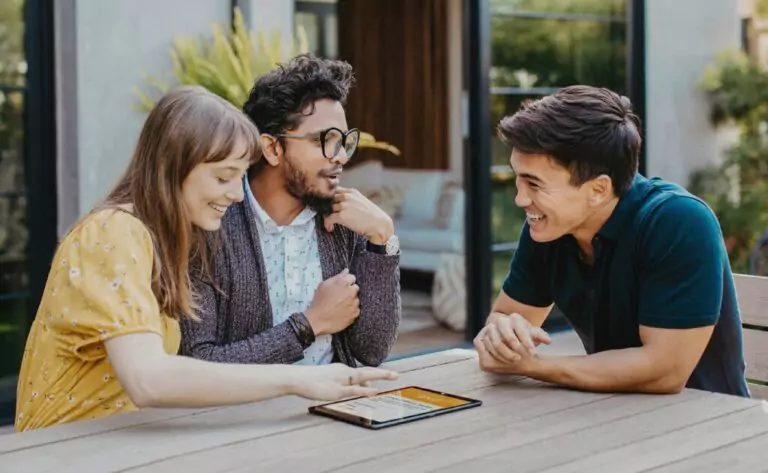 Three people sitting at a table outside, smiling and discussing finance options while looking at a tablet.