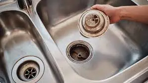 A Hand Lifts A Metal Sink Strainer From A Kitchen Sink Drain, Showing The Opening And Plumbing Inside.