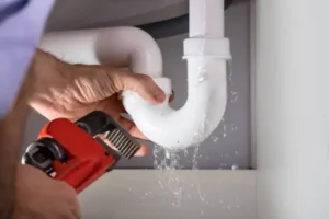 A Person Uses A Pipe Wrench To Fix A Leaking White Plastic Pipe Under A Sink, With Water Dripping From The Joint.